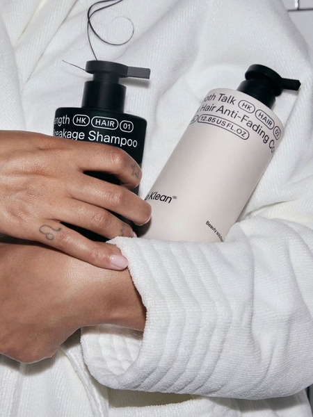 A close-up, high-contrast shot of a person in a white robe holding two minimalist hair care bottles, emphasizing brand aesthetic and product clarity.