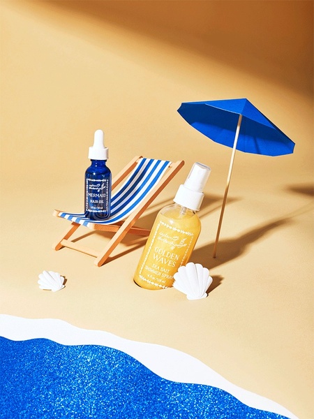 A miniature beach set featuring hair oil and shimmer spray bottles, positioned on a sand-colored surface with a small deck chair and parasol.