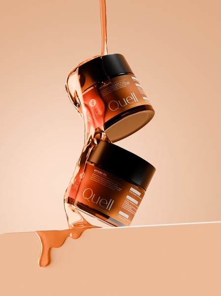 A high-end, minimalist product shot featuring two jars of Batana Oil stacked with a continuous, fluid pour of oil connecting them against a warm, neutral backdrop.