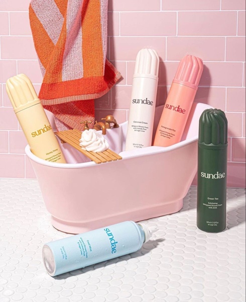 A visually striking product photography shot of multiple whipped shower foam bottles arranged around a miniature pink bathtub.
