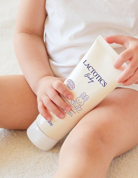 A close-up studio shot of a baby holding a tube of Lactotics Baby moisturizer, emphasizing the product's natural and gentle branding.