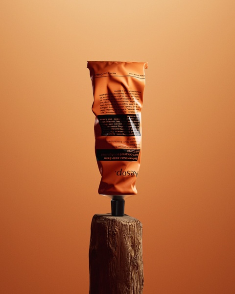 An orange body balm tube is balanced perfectly upright on a wooden post against a matching monochromatic orange background.