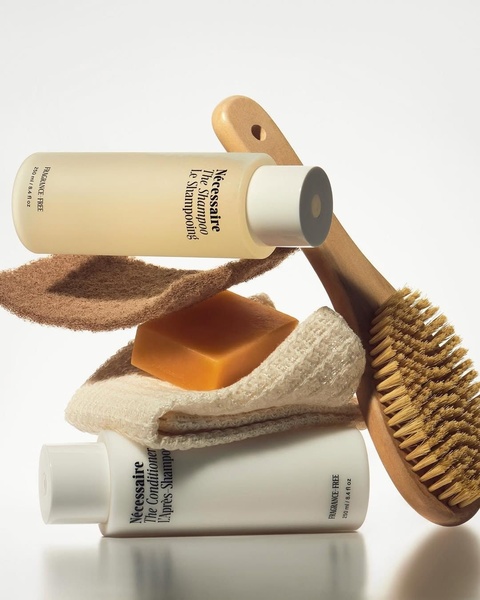 A clean, high-key studio photograph featuring bottles of shampoo and conditioner stacked with a wooden bath brush and texture pads.