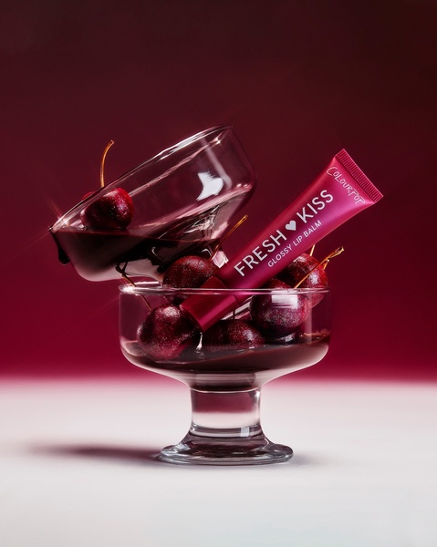 A high-fashion still life shot of a glossy lip balm tube nestled in a cocktail glass filled with fresh cherries and dark liquid.