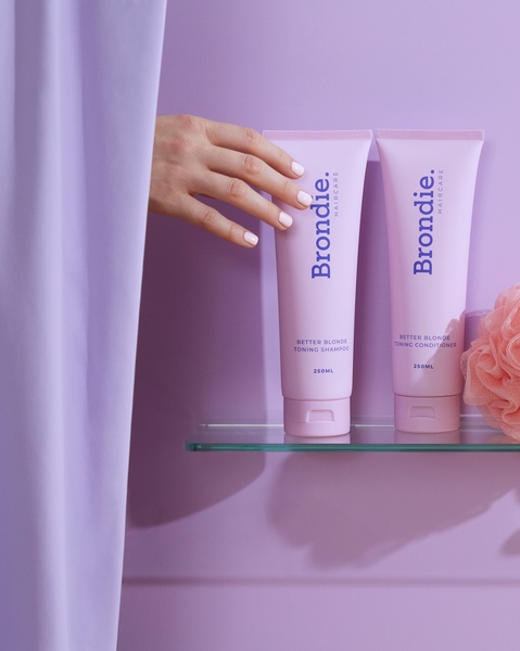 A clean, high-contrast product photography shot of shampoo and conditioner on a shelf with a soft pastel purple background and a natural hand placement.