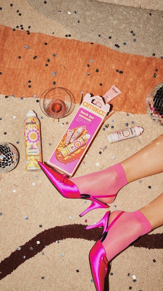 A high-angle product shot of dry shampoo bottles arranged with party props and high-heeled shoes on a textured rug.