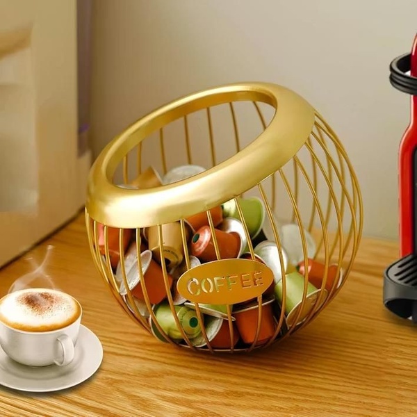 A high-end, clean product shot of a spherical gold wire coffee pod holder placed on a wooden surface next to a freshly brewed cup of coffee.