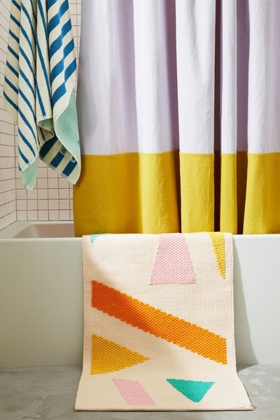 A high-angle product shot of a textured bath mat with geometric patterns, placed in front of a clean, coordinated shower curtain and towel set.