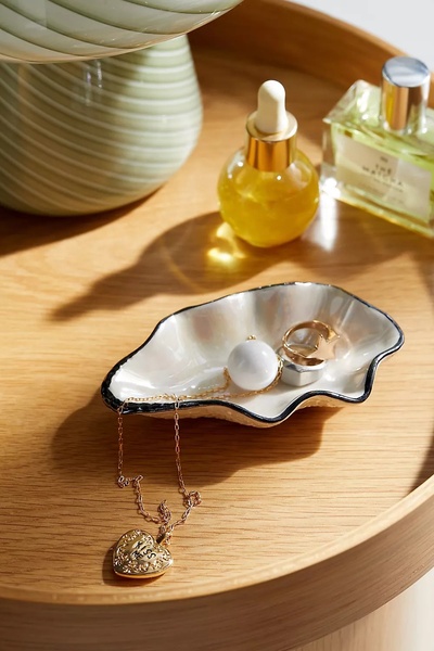 A close-up, top-down shot of a shell-shaped jewelry tray holding a gold necklace and ring, set on a wooden tray with a bottle of matcha fragrance nearby.