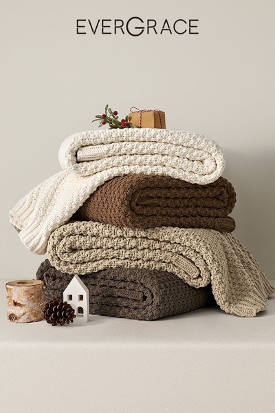 A clean, minimalist product hero shot featuring a vertical stack of knit blankets in neutral tones with seasonal holiday accents.