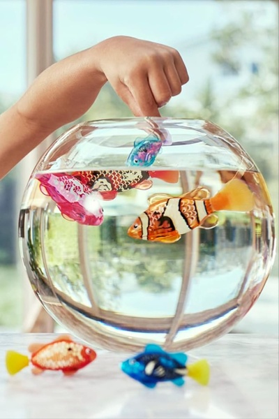 A close-up shot showing a child interacting with robotic toy fish swimming in a clear glass bowl, emphasizing movement and tactile play.