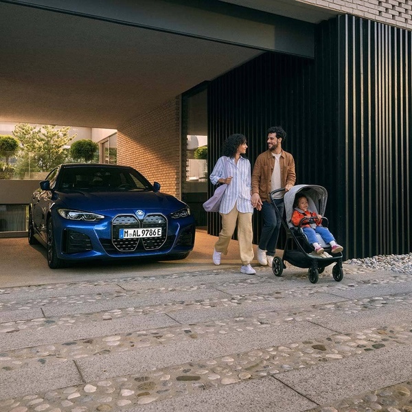 A high-quality lifestyle image of a young couple with a baby stroller posing next to a blue electric sedan parked in a modern home driveway.