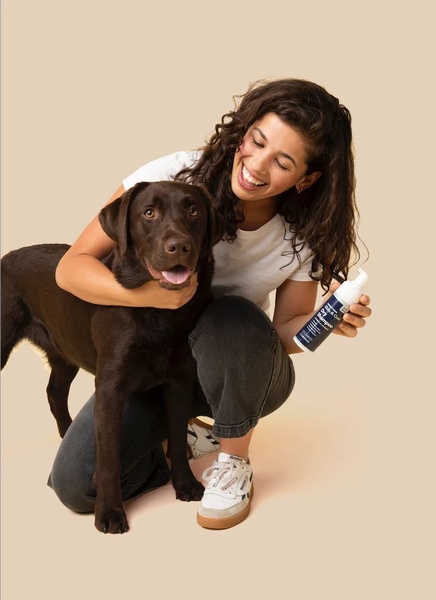 A woman happily hugs her chocolate Labrador while holding a bottle of dog dry shampoo, creating an authentic, relatable moment for pet owners.