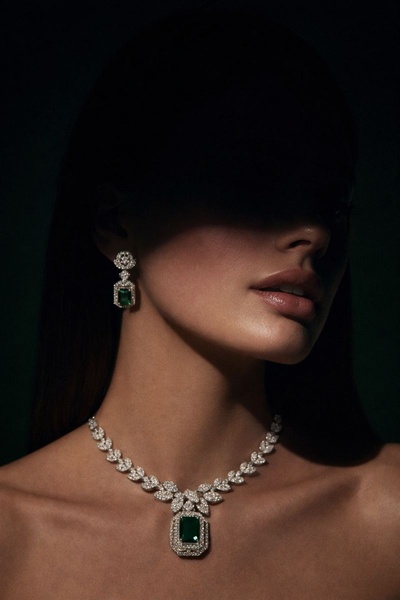 A high-end portrait featuring a model wearing a luxurious emerald and diamond necklace with matching earrings against a dark, moody background.