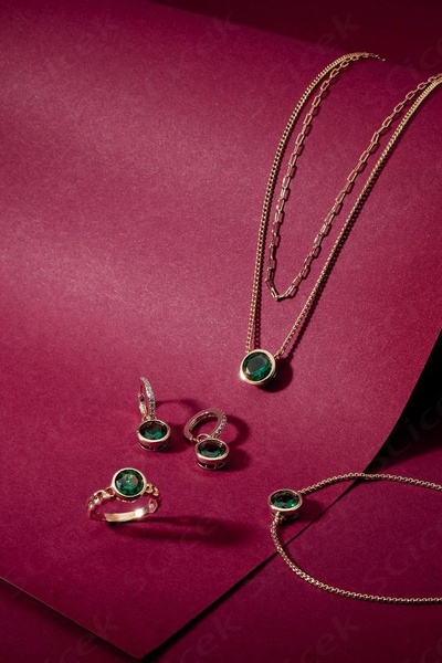 A sophisticated, professional studio product shot featuring a matching emerald necklace, ring, and earrings set against a deep burgundy backdrop.