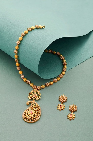 A high-end, clean product shot of a gold necklace set with embedded stones, displayed against a curved, soft-toned background.