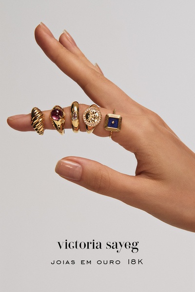 A close-up, high-fashion shot of a hand wearing five distinct, elegant gold rings against a neutral, minimalist backdrop.