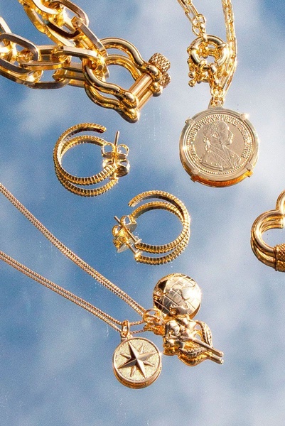 A high-end, reflective flat-lay shot of gold chains, coin pendants, and hoop earrings against a blue sky background.