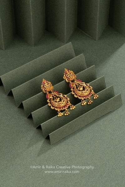 A refined product close-up featuring gold earrings with ruby accents, set on an accordion-folded textured green background.
