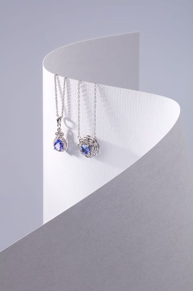 A high-end, studio-quality photograph featuring two delicate silver necklaces with blue gemstone pendants draped elegantly over a curved white paper backdrop.