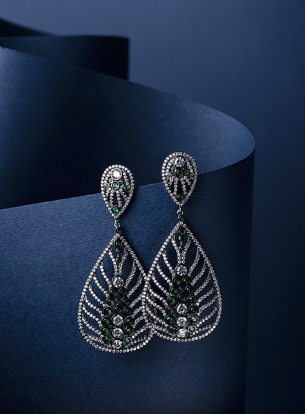 A high-end studio capture of ornate emerald and crystal earrings resting on a curved deep blue paper background.