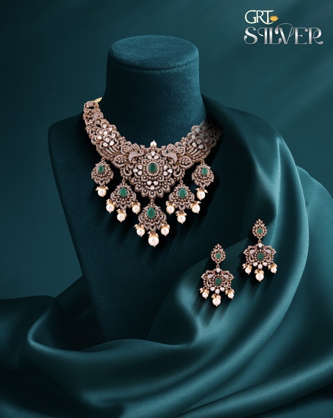 A high-end, studio-lit hero shot of a silver necklace with emeralds and pearls, set against a deep teal silk drape.