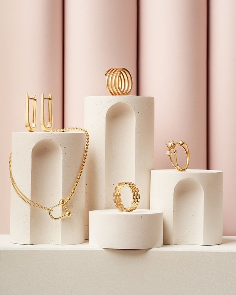 A high-end, clean-aesthetic studio shot featuring various gold jewelry pieces arranged on textured white pedestals against a soft pink curved background.