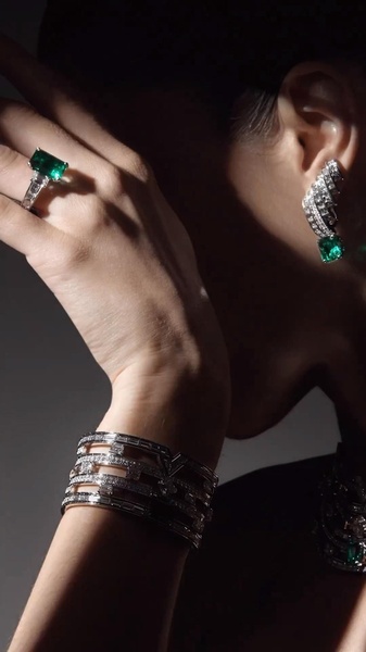 A sophisticated close-up shot focusing on a high-value emerald and diamond ring paired with matching earrings and bracelet in a dimly lit, moody environment.