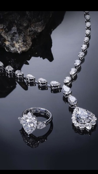 A high-contrast studio shot featuring a pear-cut diamond necklace and matching ring against a dark, reflective surface.
