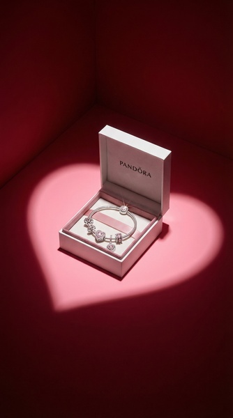 A high-contrast studio shot of a charm bracelet inside an open jewelry box, dramatically illuminated by a heart-shaped spotlight.