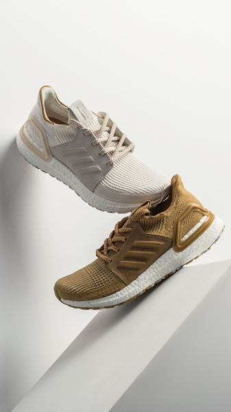 A high-contrast vertical shot featuring two running shoes in neutral earth tones suspended against a clean white geometric backdrop.