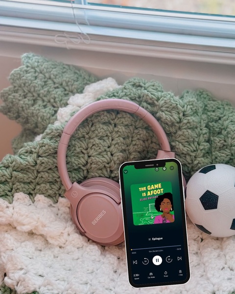 A close-up aesthetic shot of pink headphones next to a phone displaying a podcast, all set against a soft, knitted green and white textured background with a soccer ball.