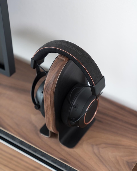 A clean, high-end product display showing premium headphones resting on a custom wooden and metal stand. The composition uses neutral tones and soft lighting to emphasize craftsmanship.