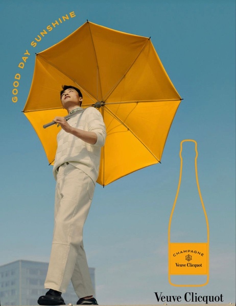 A high-contrast, lifestyle-focused ad for champagne featuring a man holding a bright yellow umbrella against a blue sky, conveying warmth and premium optimism.