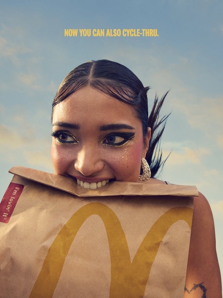 A close-up portrait of a woman holding a branded paper bag in her teeth against a blue sky, framing a casual, youthful lifestyle.