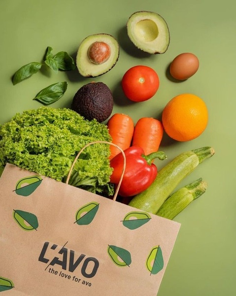 A top-down view of a L'AVO branded shopping bag overflowing with fresh produce like avocados, tomatoes, and vegetables against a solid green background.