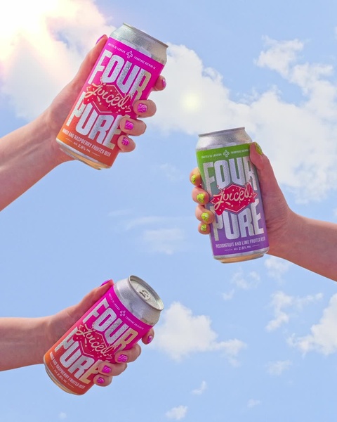A vibrant, low-angle shot showing three hands holding cans of fruited beer against a bright blue sky to evoke refreshment and summer fun.