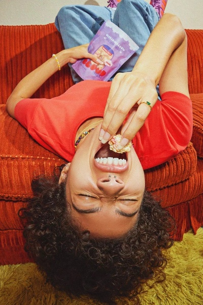A casual, top-down shot of a user laying on an orange couch, eating marshmallows directly from the bag in a relatable, fun, and unpolished style.