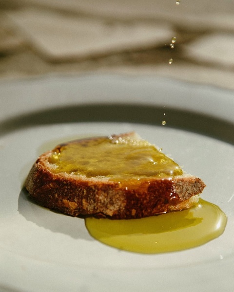 A close-up, high-contrast shot of golden olive oil being drizzled over a slice of rustic bread on a simple ceramic plate.