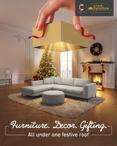 A creative showing a giant hand placing a gift box over a modern living room setting with a sectional sofa, Christmas tree, and cozy fireplace.