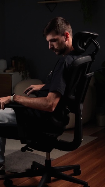 A high-contrast, moody shot featuring a man sitting in an ergonomic office chair in a dimly lit room, highlighting the product design and comfort.