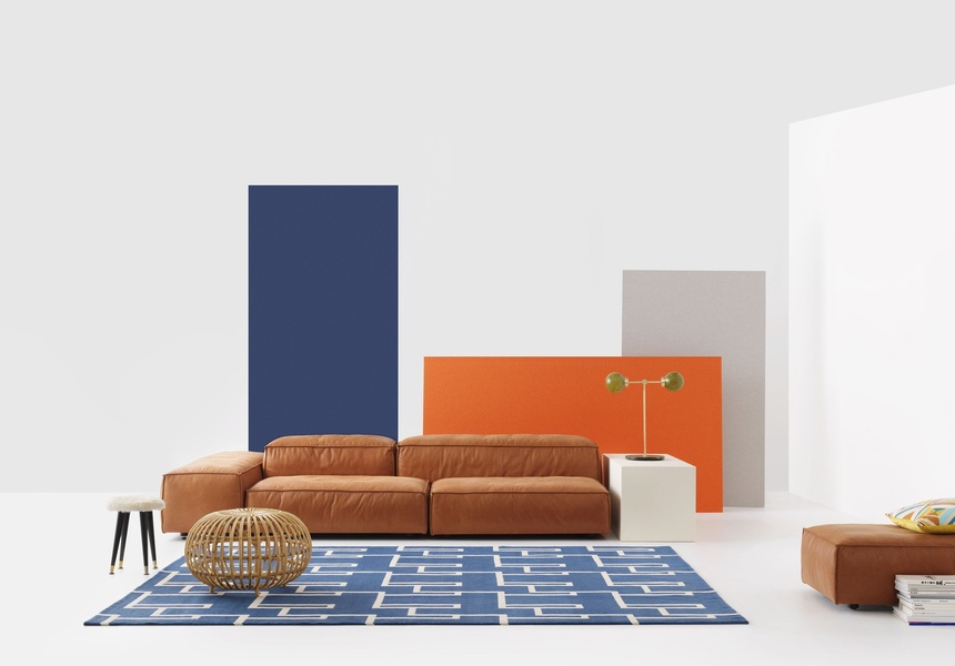 A high-end leather sofa is centered against a clean white wall, framed by primary-colored geometric panels that define the product's premium aesthetic.