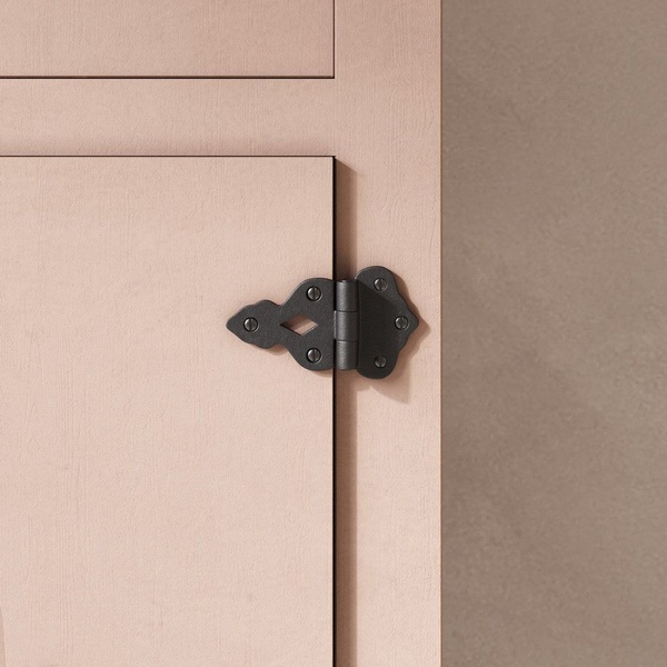 A high-detail close-up of a matte black ornate cabinet hinge installed on a muted pink cabinet door, focusing on product quality and texture.