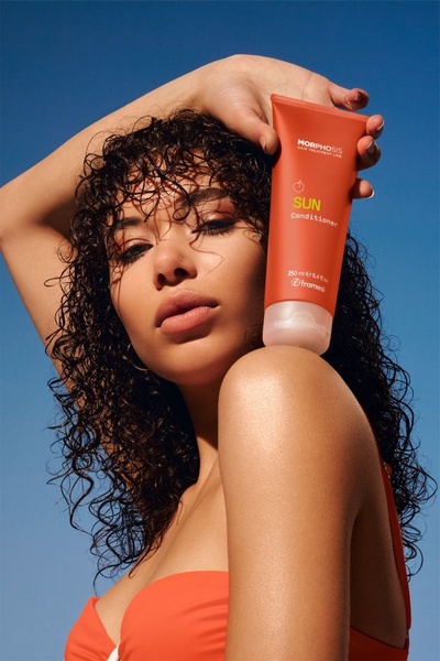 A close-up portrait of a woman holding a sun-care hair product against a clear blue sky, emphasizing the summer season.
