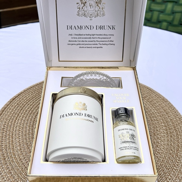 A high-end, top-down product hero shot displaying a jewelry cleaner kit in a luxury gold-trimmed open box on a textured mat.