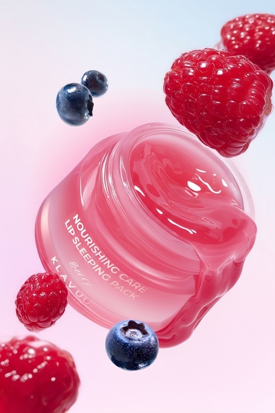 A high-quality studio shot featuring a lip sleeping pack jar surrounded by floating raspberries and blueberries, with product texture oozing from the open lid.