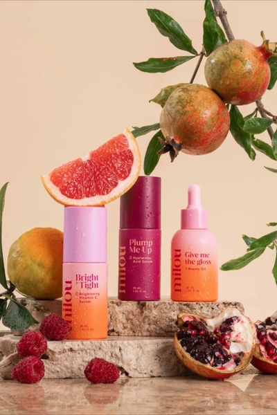 This creative showcases three distinct skincare serums arranged on natural stone against a soft beige background, surrounded by fresh pomegranate and citrus fruit.