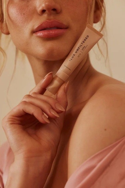 A close-up, soft-focus shot of a model holding a tube of lip serum against a neutral background, highlighting natural beauty and product texture.