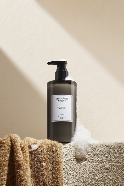 A high-end studio product photograph featuring a shampoo bottle resting on textured stone with soap foam and a neutral-toned towel.