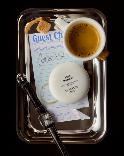 A high-end product photo featuring hand care cream on a restaurant tray with coffee and a wristwatch.
